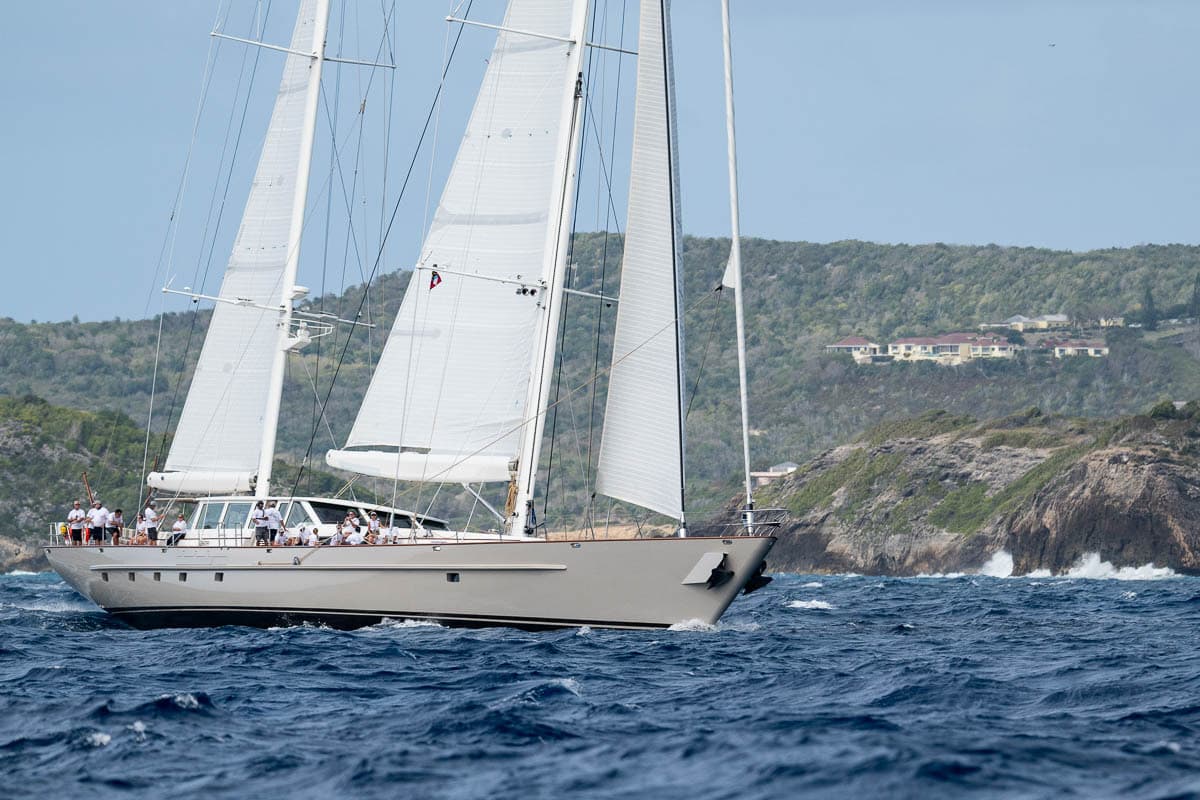 Sailing yacht Catalina competing at the Antigua Superyacht Challenge 2025, sailing close to the coastline with crew members dressed in white shirts, rugged cliffs and buildings visible in the background.