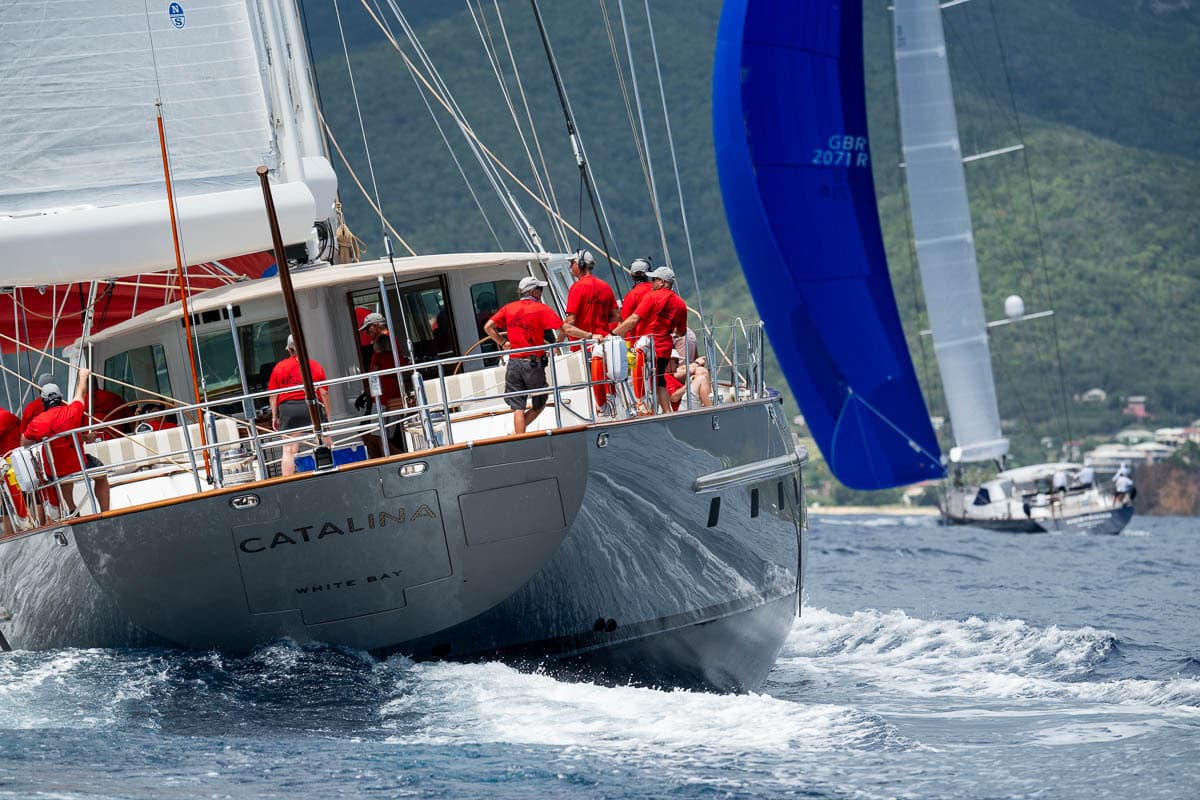 Sailing yacht Catalina competing in the Antigua Superyacht Challenge 2025, crew in red shirts working on deck as the yacht heels over on the water with another yacht racing in the background.