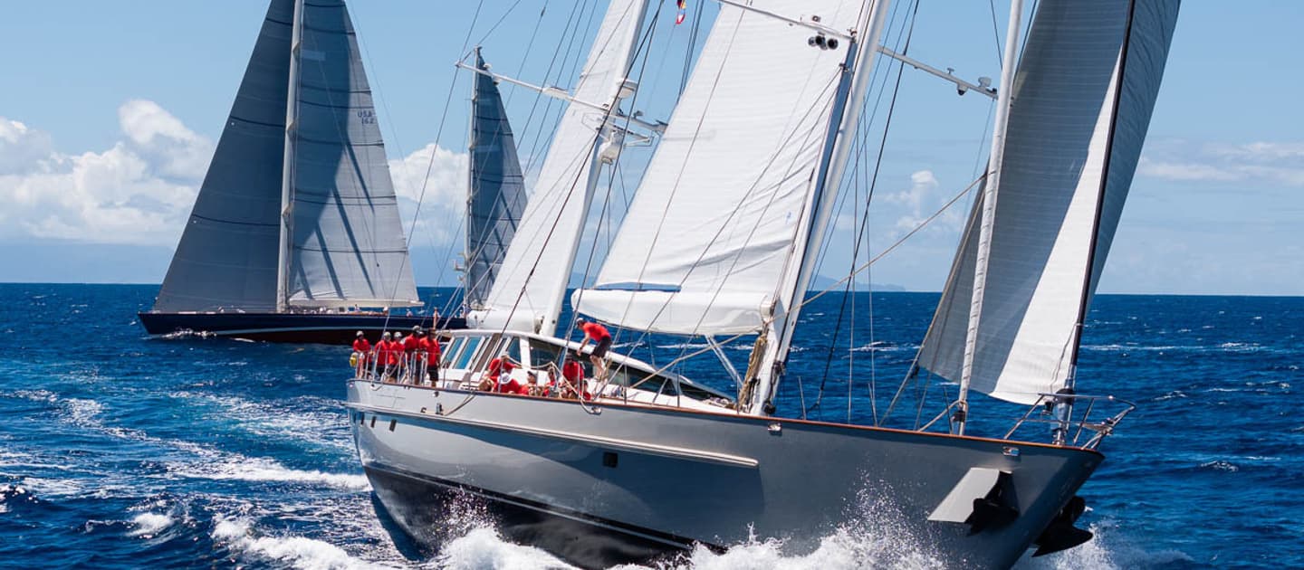 Sailing yacht Catalina racing at the Antigua Superyacht Challenge 2025, crew in red shirts adjusting sails as the yacht heels through the waves, with a yacht visible in the distance.