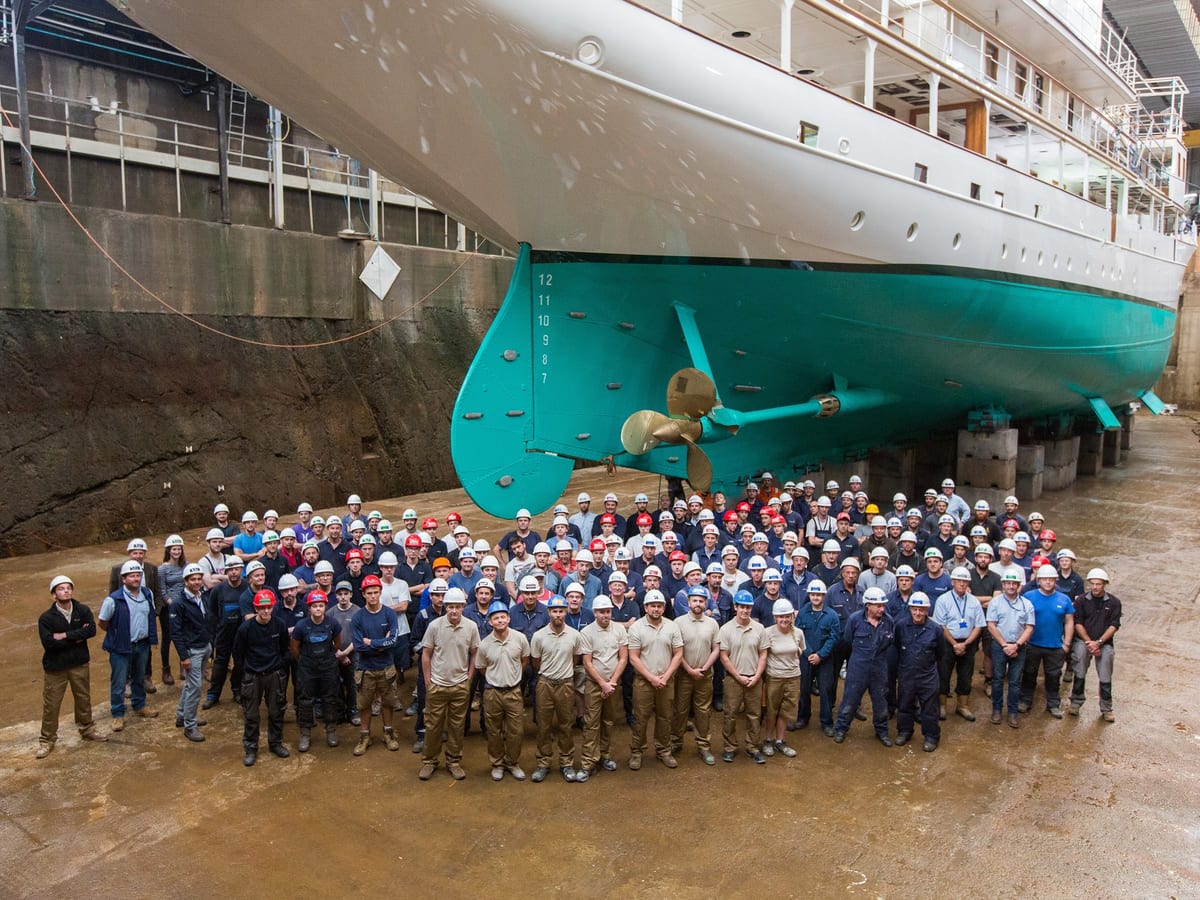 Pendennis team standing beneath Haida 1929 in dry dock, with her freshly painted turquoise hull and golden propeller.