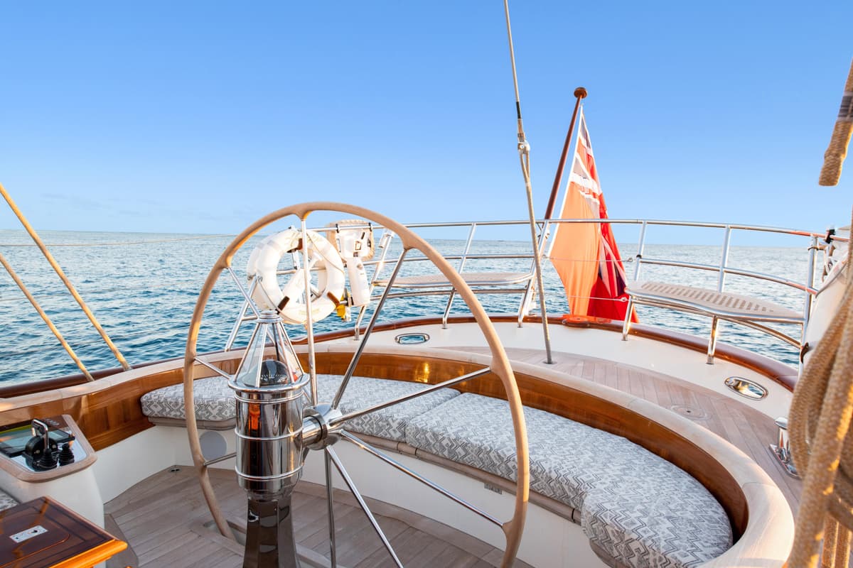 Helm station aboard Halekai with polished wheel, curved bench seating, and Red Ensign flag at sea.