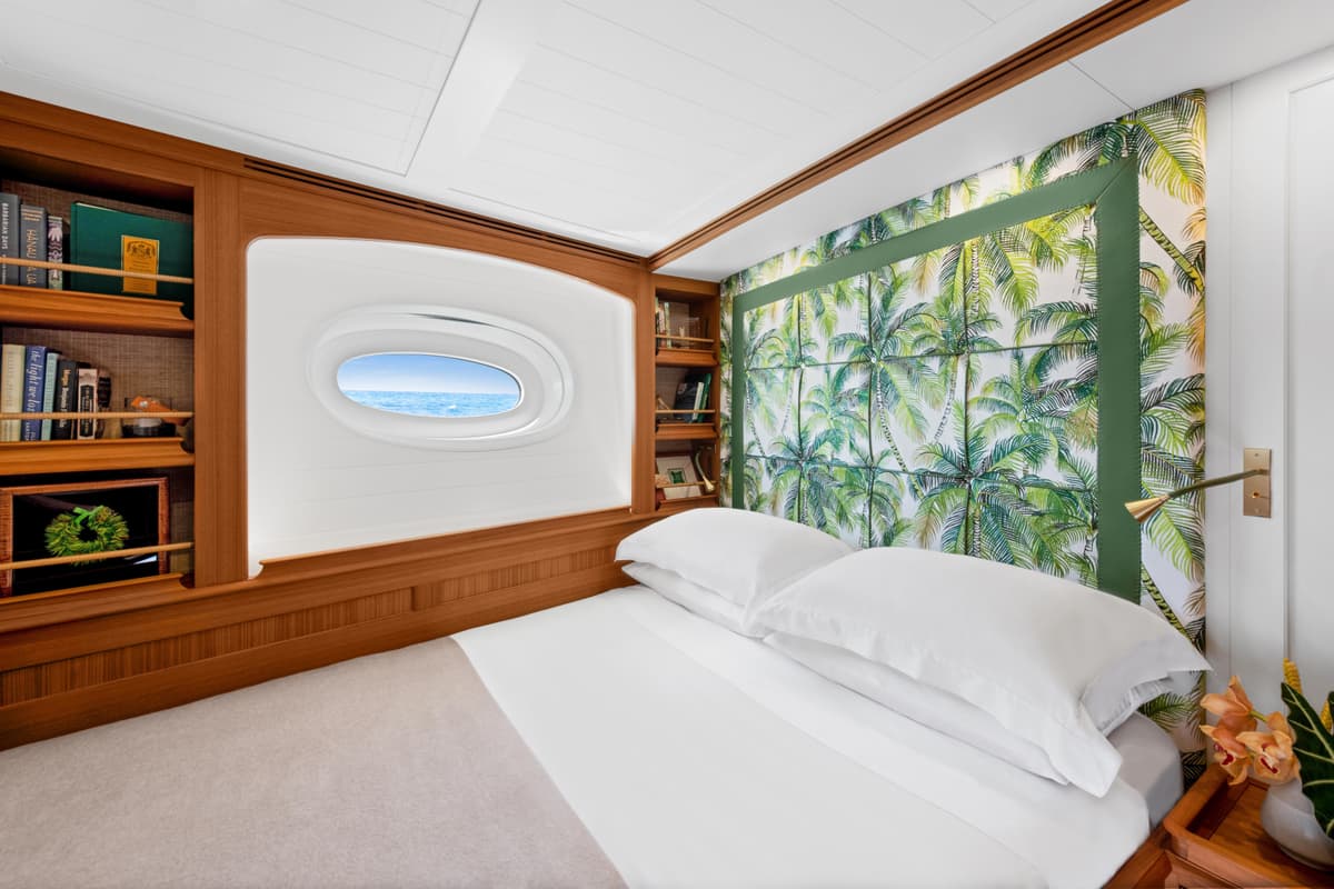 Halekai guest cabin with tropical palm-print headboard, porthole sea view, and warm timber cabinetry.
