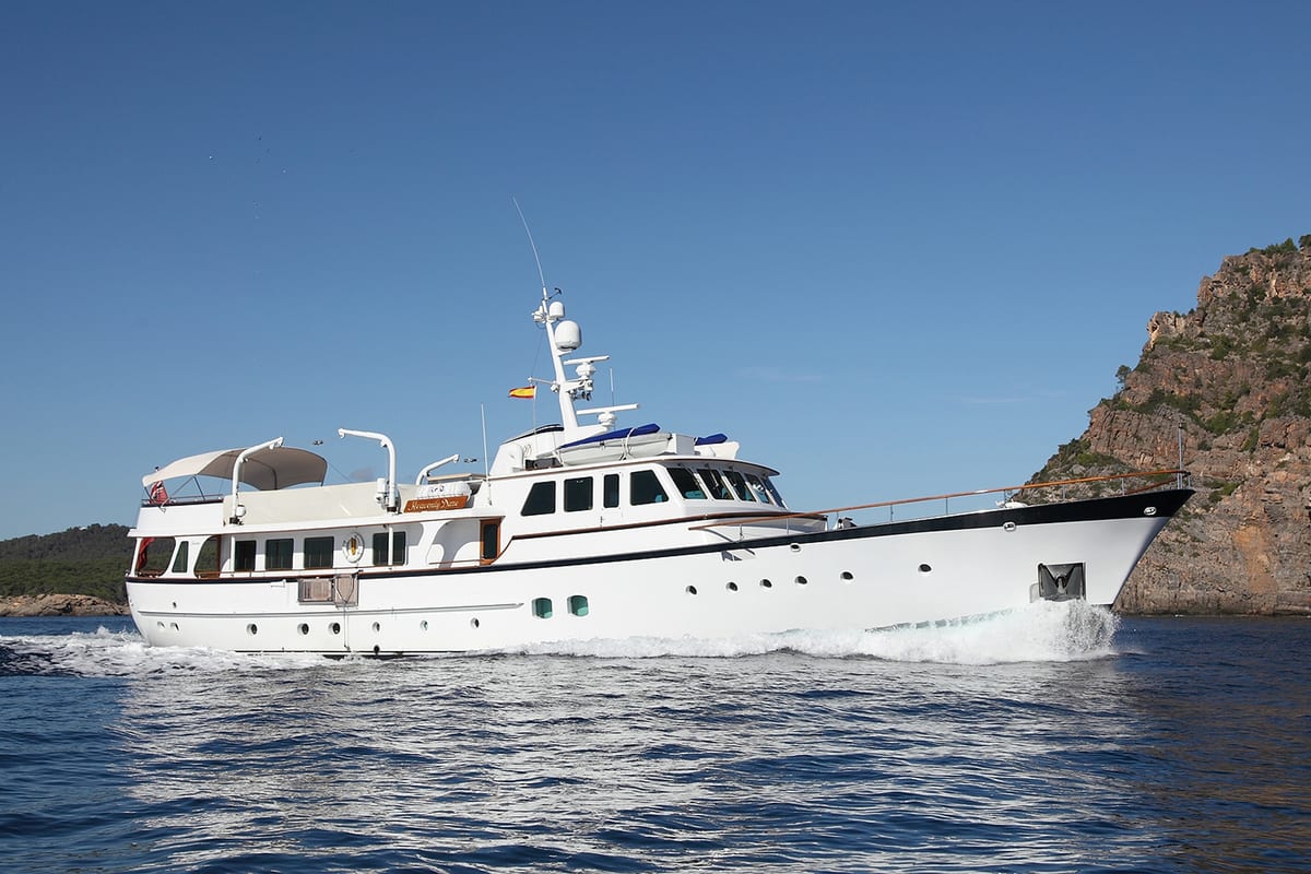 Classic motor yacht Heavenly Daze cruising along a rocky Mediterranean coastline under clear blue skies.