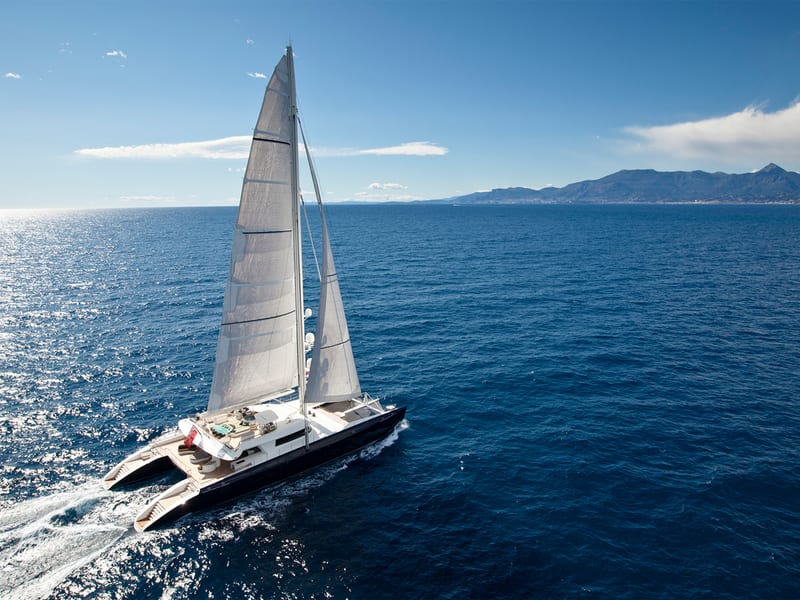 Sailing catamaran Hemisphere cruising in open water with coastline and mountains in the distance.