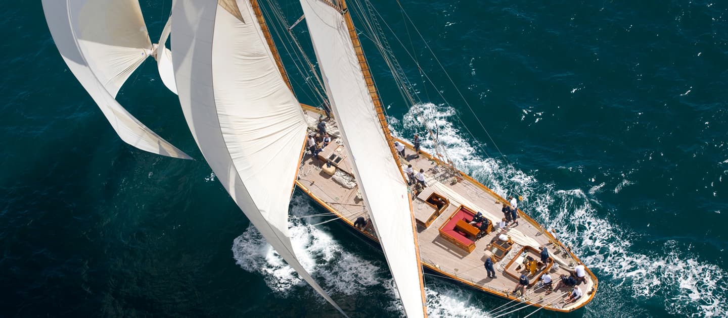 Aerial view of Mariette under full sail with crew on deck, racing through open water.