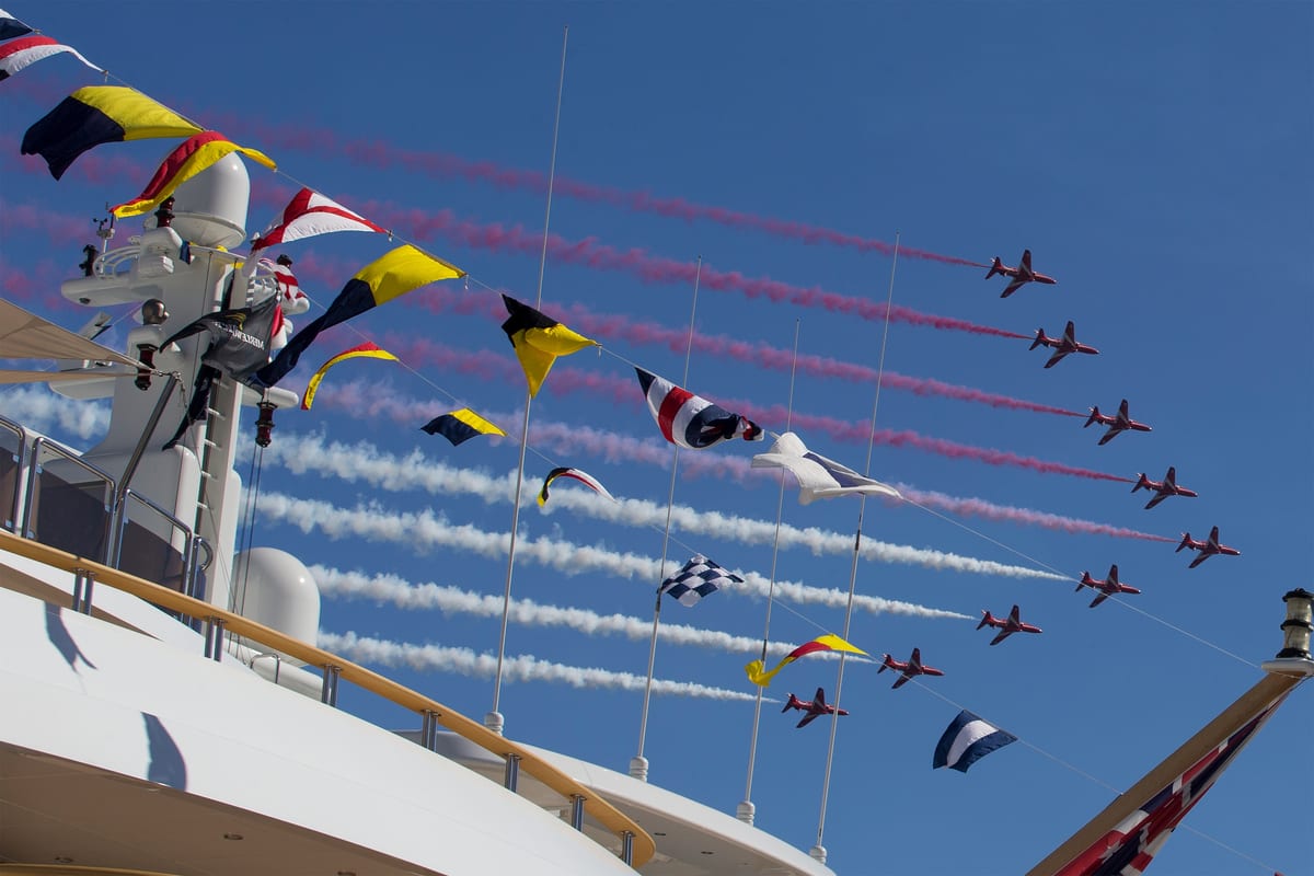 Red Arrows fly in formation above a superyacht deck adorned with colourful signal flags, trailing red, white, and blue smoke.