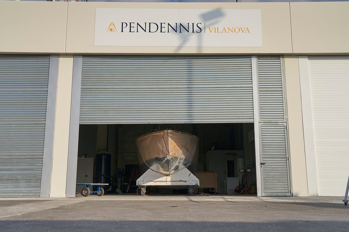 Wooden yacht hull under restoration inside the Pendennis Vilanova facility, viewed through a partially open workshop shutter.