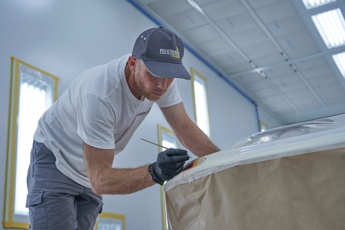 Pendennis Vilanova team member hand-painting detail work on a masked yacht surface inside a brightly lit workshop.