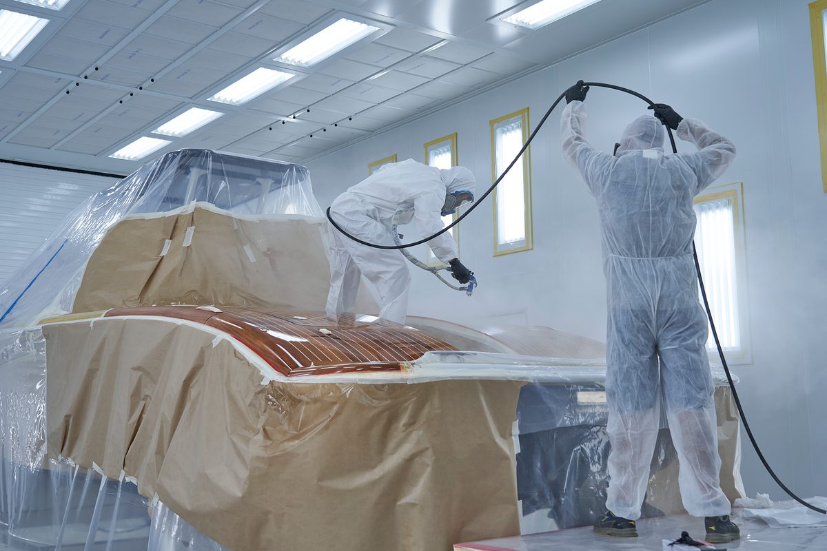 Two Pendennis Vilanova technicians in protective suits spray paint a masked yacht structure inside a dedicated climate-controlled facility.