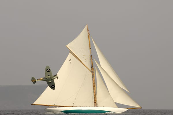 A Spitfire flies low past a classic gaff-rigged yacht with full sails during the 2010 Pendennis Cup.