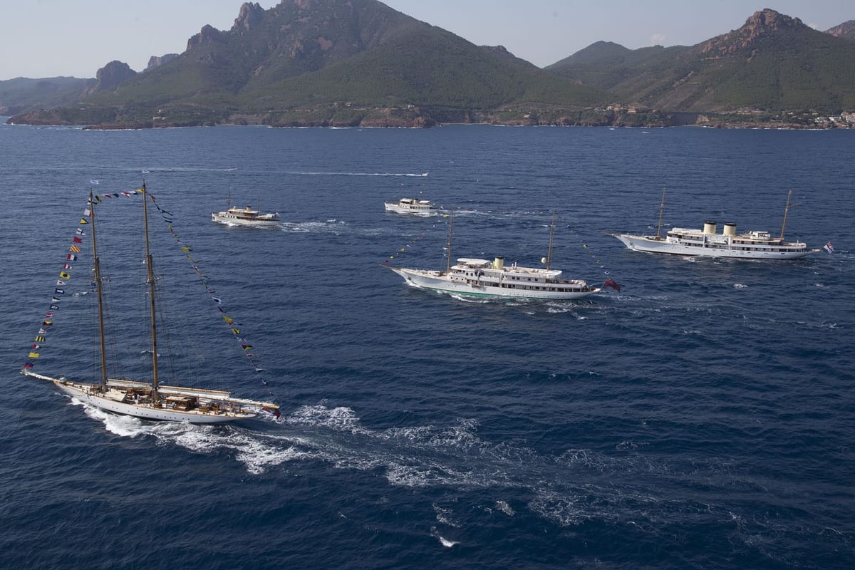 Classic schooner and motor yachts cruise in formation near a rugged coastline, with signal flags fluttering in the breeze.