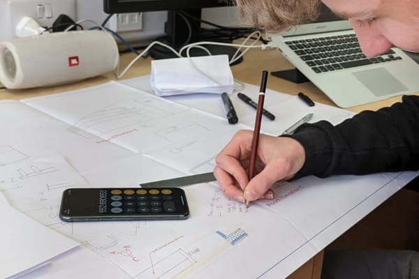 Apprentice marks measurements on a race control room plan at a desk, with calculator and laptop nearby.