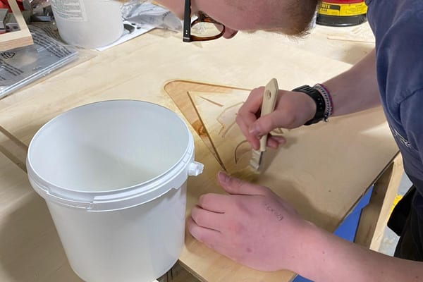 Apprentice carefully paints a Flushing Sailing Club burgee design onto a wooden panel as part of race furniture detailing.