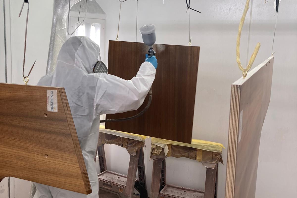 Apprentice in protective gear spray-finishes wooden panels in a booth during construction of Flushing Sailing Club race furniture.