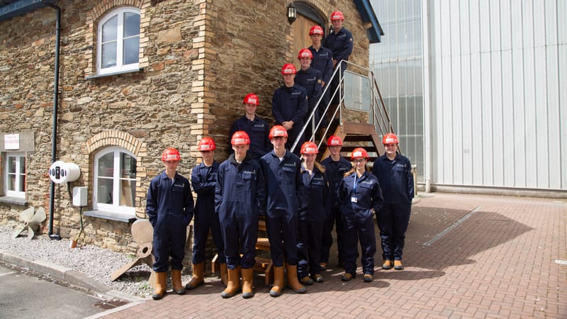 Group of Pendennis apprentices in hard hats and overalls outside a stone building at the Falmouth shipyard.