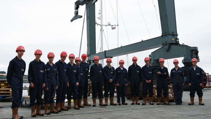 Pendennis apprentices in overalls and red hard hats standing in front of the shipyard's lifting gantry, marking the 200th apprentice milestone.