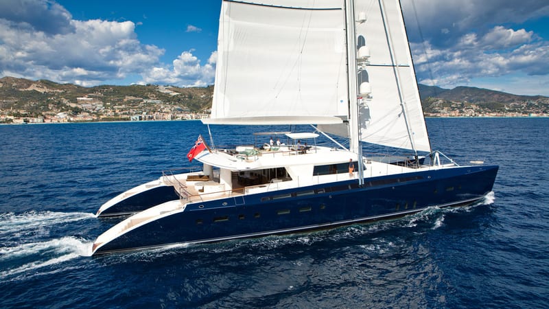Luxury catamaran Hemisphere sailing under full sail in deep blue waters near a coastal town, showcasing Pendennis’s custom build expertise.