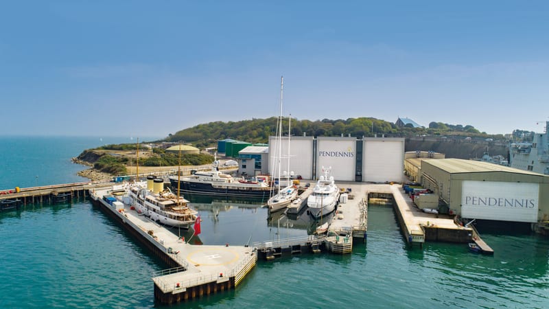 Aerial view of Pendennis Shipyard's non-tidal wet basin with several superyachts docked and shipyard buildings in the background.