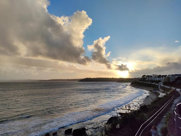 Dramatic sunset over Gyllyngvase Beach in Falmouth, with waves rolling in and a golden glow breaking through the clouds.