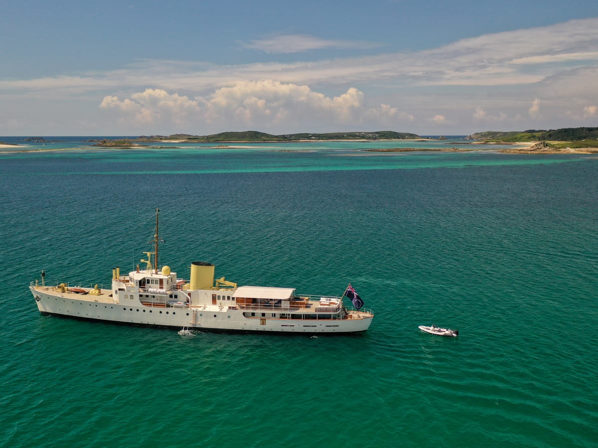 Classic motor yacht Marala anchored in the turquoise waters off the Isles of Scilly, with a tender alongside and sandy islands in the distance.