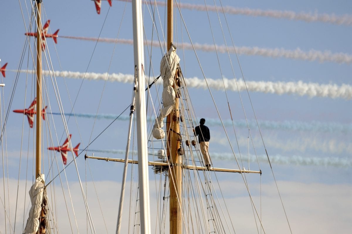 Crew member stands on the mast of a classic yacht during the 2010 Pendennis Cup, as Red Arrows jets perform overhead with colourful smoke trails.