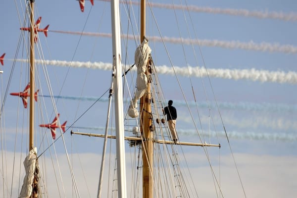 Crew member stands on the mast of a classic yacht during the 2010 Pendennis Cup, as Red Arrows jets perform overhead with colourful smoke trails.