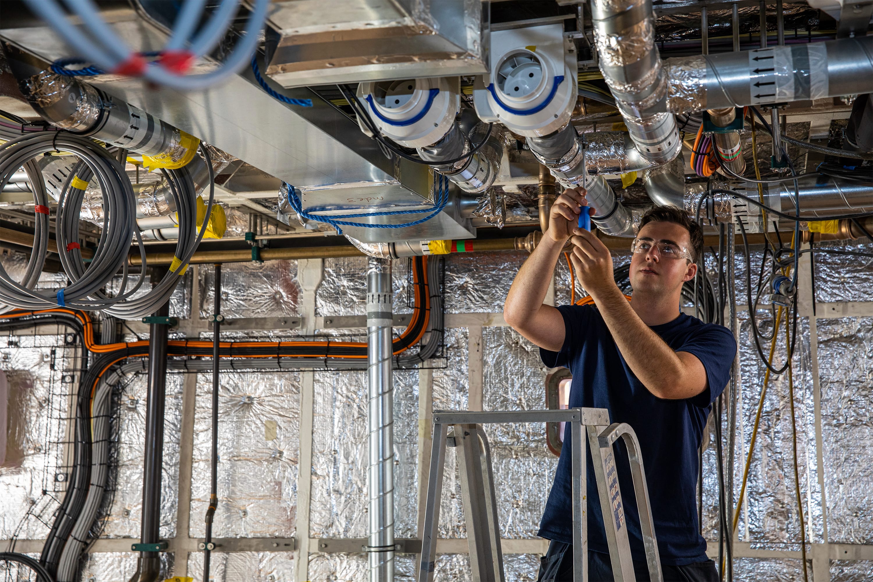 Apprentice installing overhead ductwork among exposed wiring and pipework.