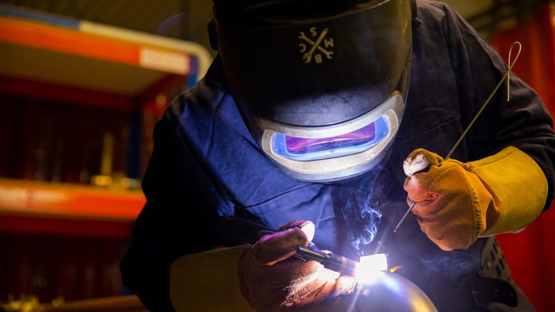 Welder in full gear focusing on a detailed weld in a workshop environment.
