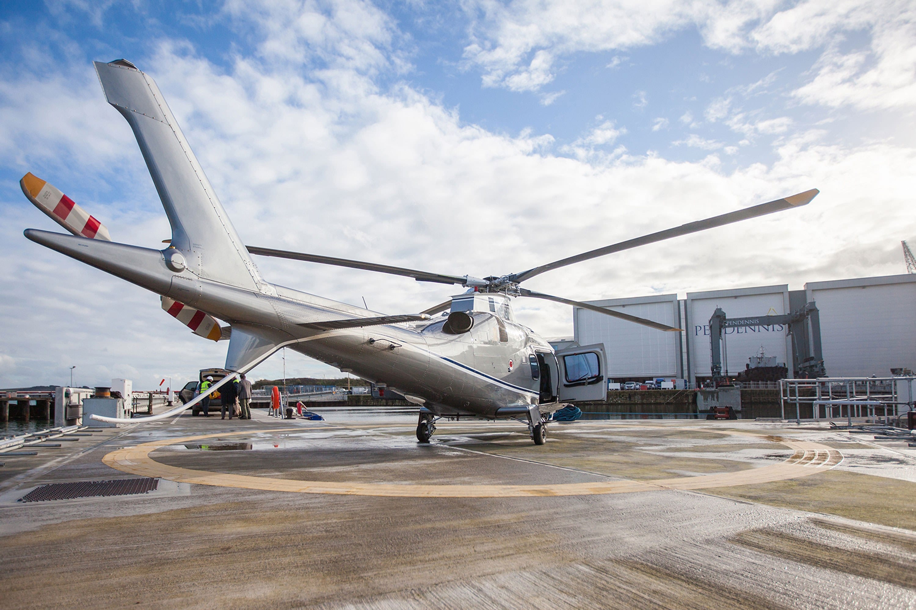 Helicopter parked on the helipad at Pendennis Shipyard, with main facility buildings visible in the background.