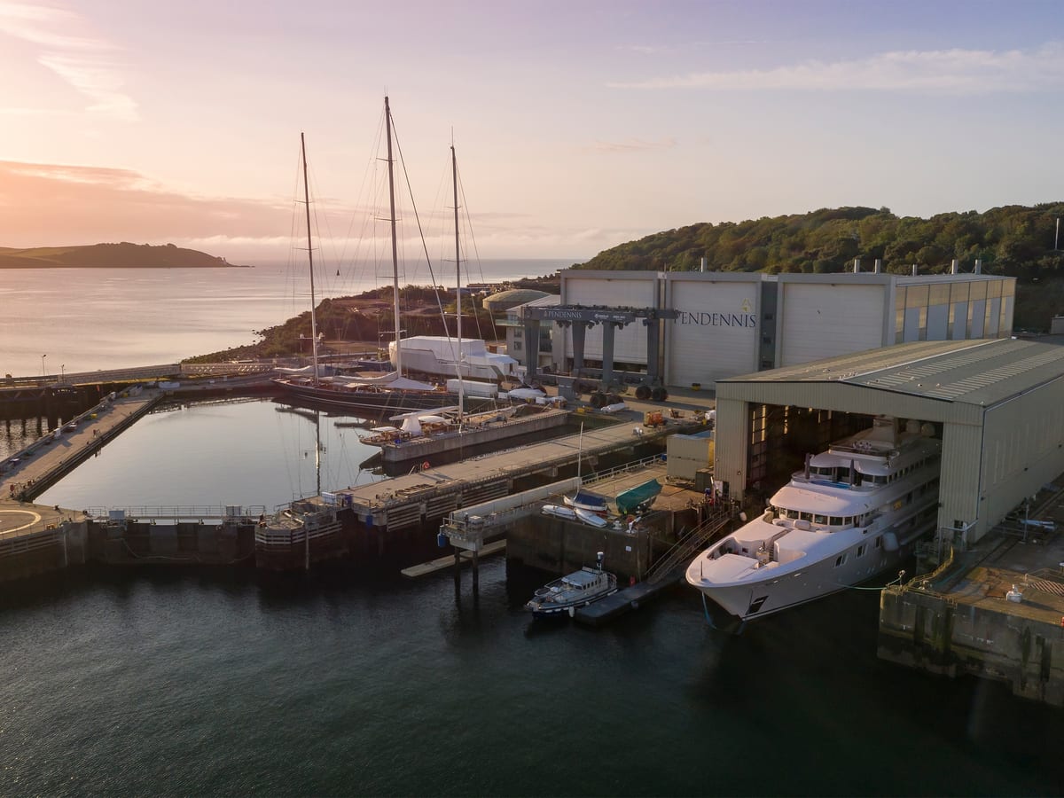 Pendennis wet basin and facilities at sunset, with superyachts docked and a motor yacht emerging from the main shed.