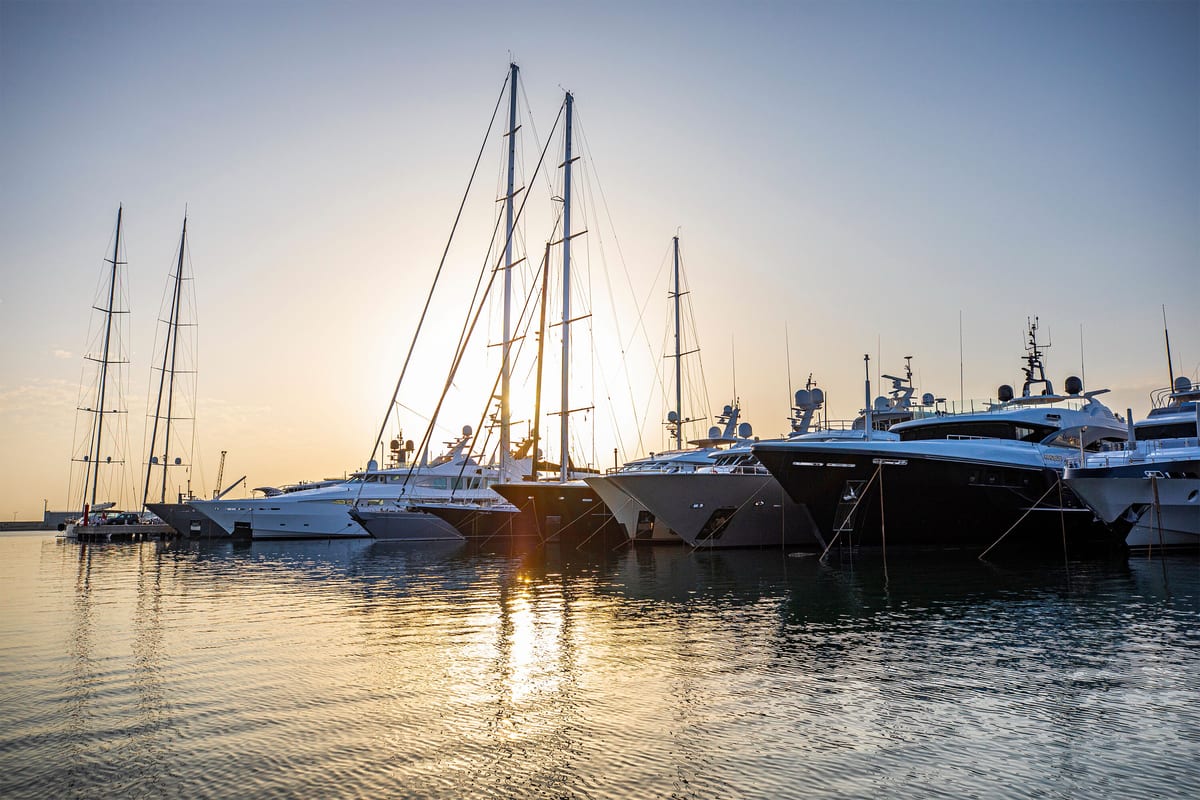 Multiple superyachts moored bow-to at Pendennis Vilanova marina during golden hour, with calm water and soft light.