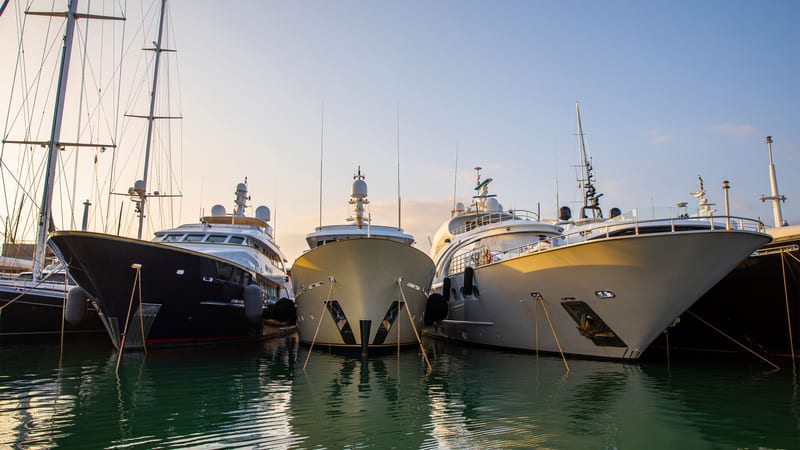 Multiple superyachts moored bow-to at Pendennis Vilanova marina during golden hour, with calm water and soft light.