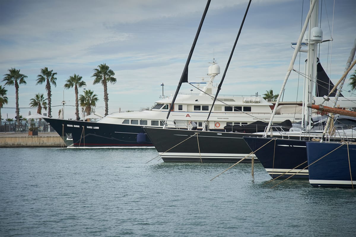 Superyachts and sailing yachts moored at Pendennis Vilanova marina, with palm-lined promenade in the background.