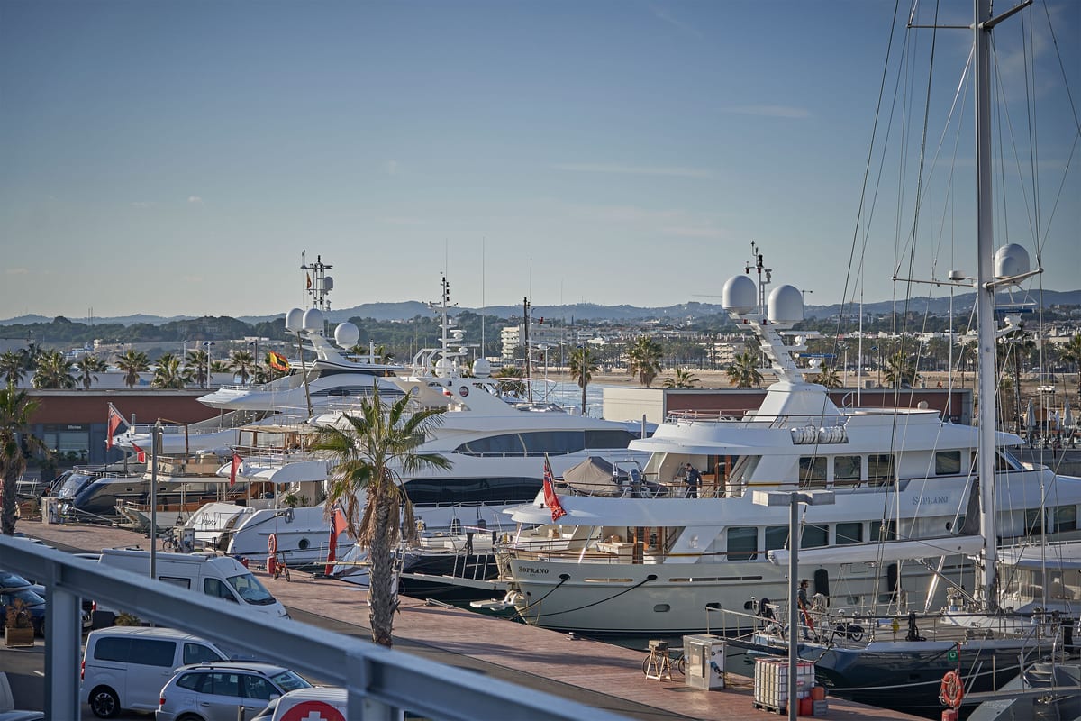 Fleet of superyachts moored at Pendennis Vilanova marina, with palm trees, waterfront buildings, and hills in the distance.