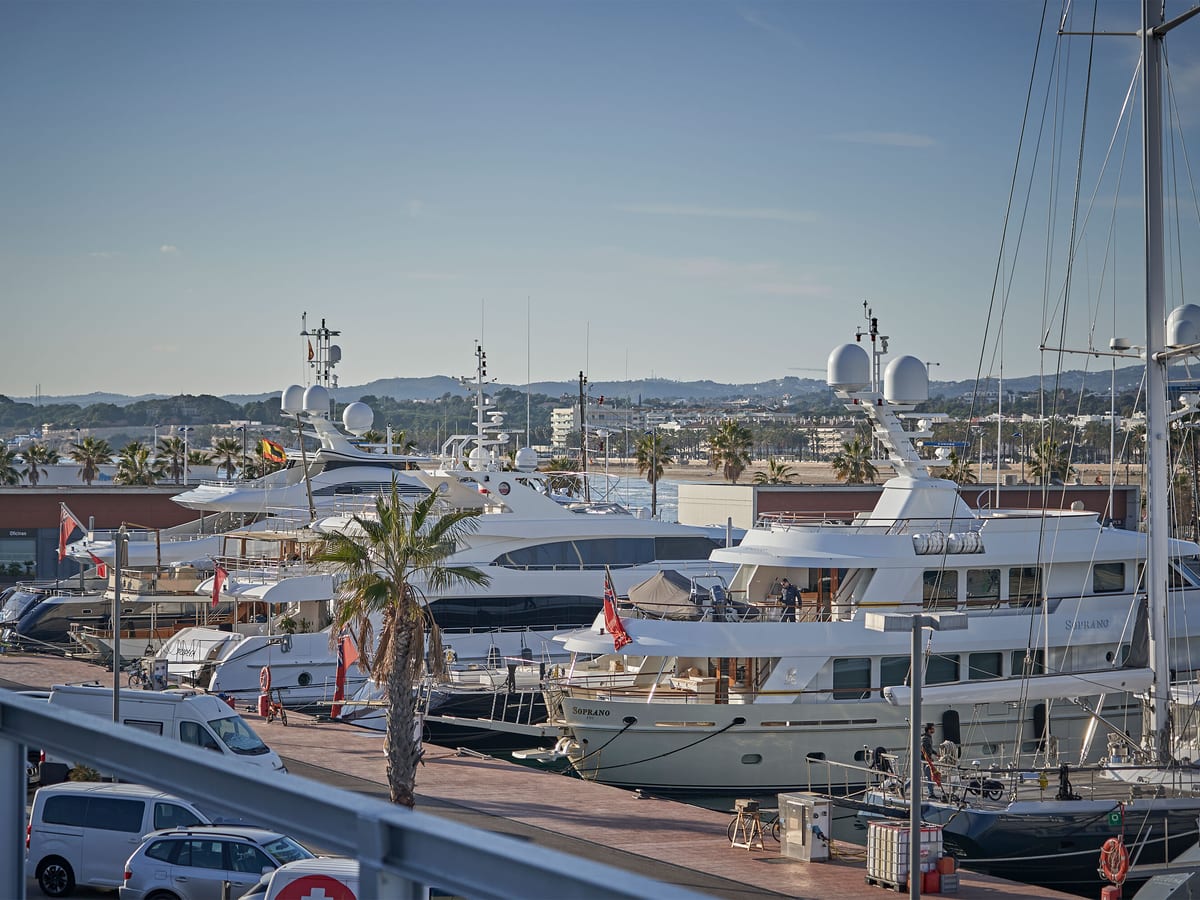 Fleet of superyachts moored at Pendennis Vilanova marina, with palm trees, waterfront buildings, and hills in the distance.
