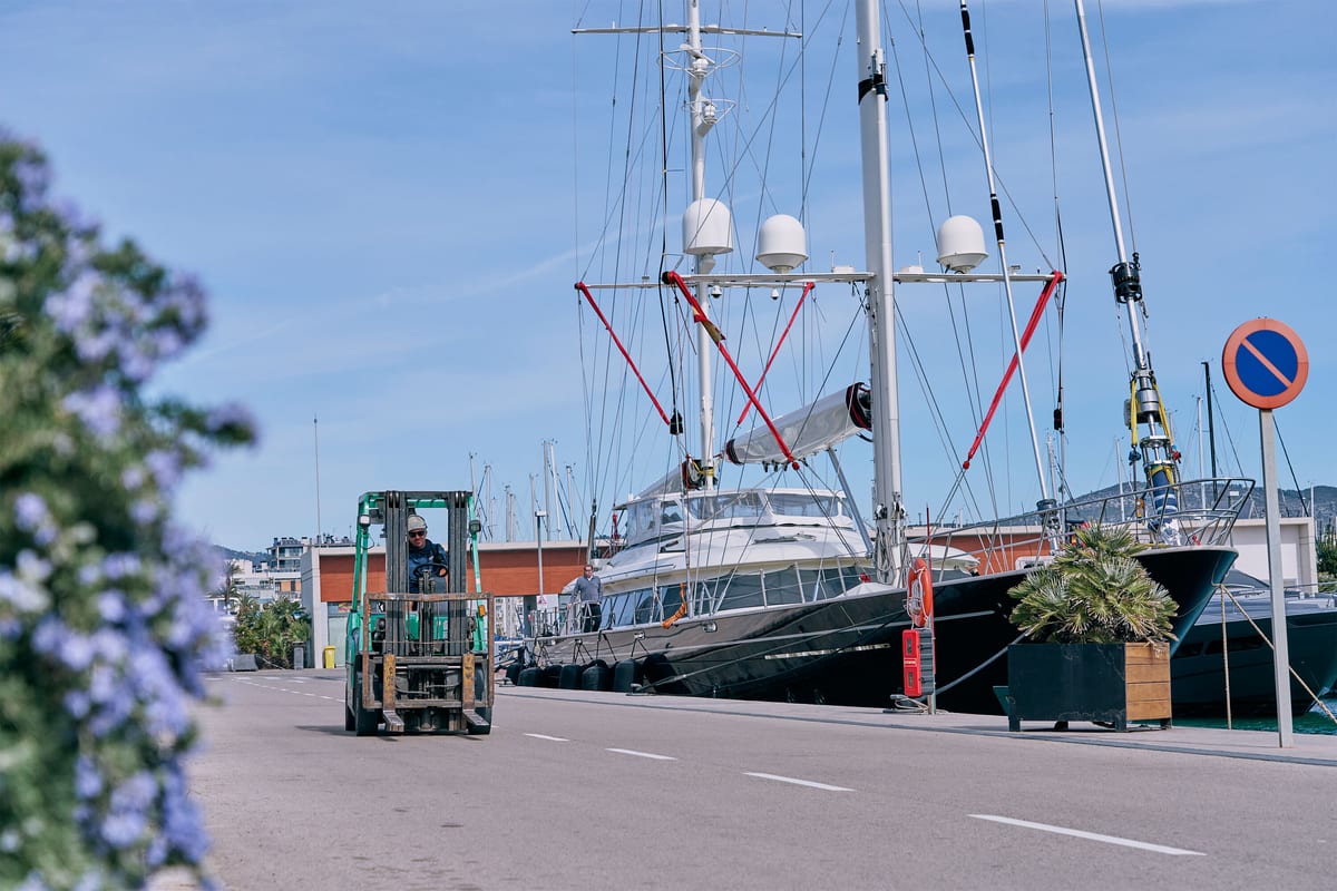 Forklift driving along Pendennis Vilanova marina with a sailing superyacht moored nearby and flowers in the foreground.
