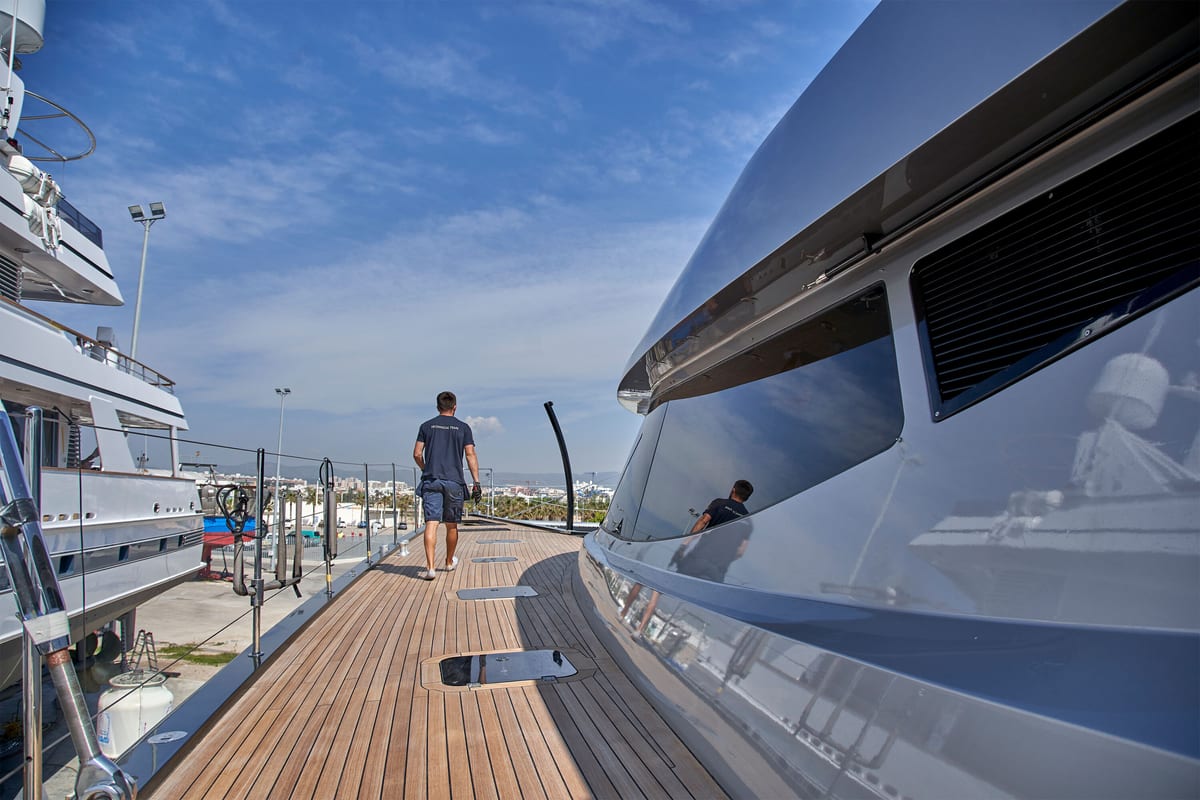 Crew member walking along the teak deck of a superyacht at Pendennis Vilanova, with reflections on the yacht’s polished hull.