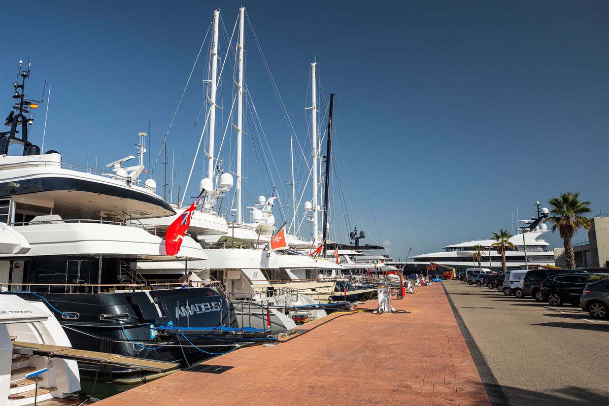 Row of superyachts berthed along the quay at Pendennis Vilanova marina, with flags flying and clear blue skies.