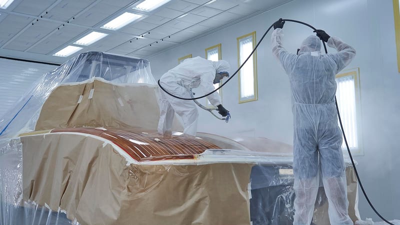 Two Pendennis technicians in protective suits spray-coating a varnished deck section inside the Vilanova paint facility.