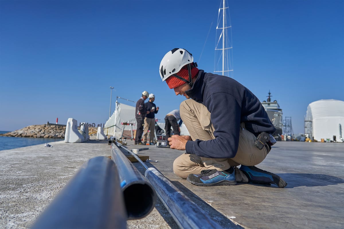 Pendennis Vilanova team member assembling metal tubing on the dockside with others working in the background.