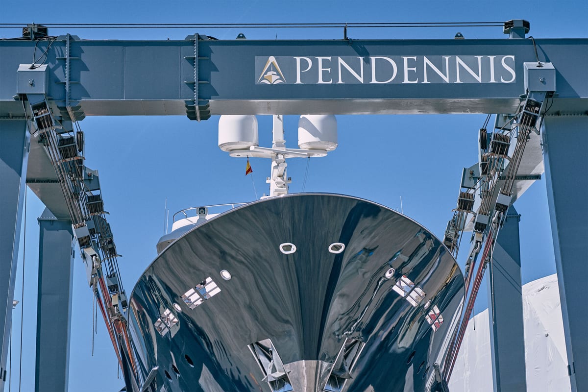 Superyacht bow suspended in Pendennis Vilanova's lifting dock beneath large blue gantry crane bearing the Pendennis logo.