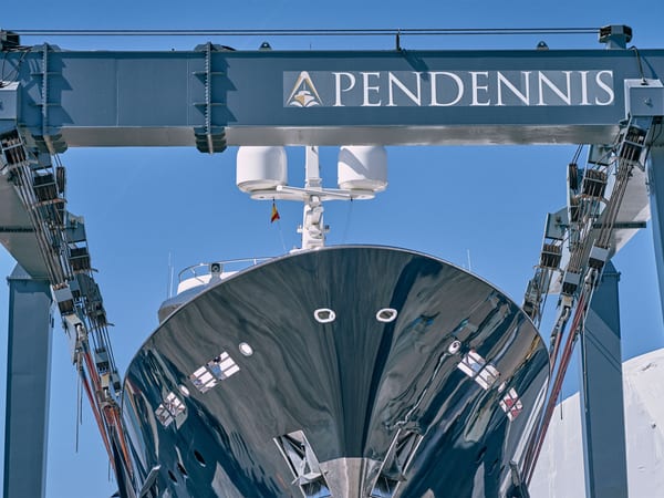 Superyacht bow suspended in Pendennis Vilanova's lifting dock beneath large blue gantry crane bearing the Pendennis logo.