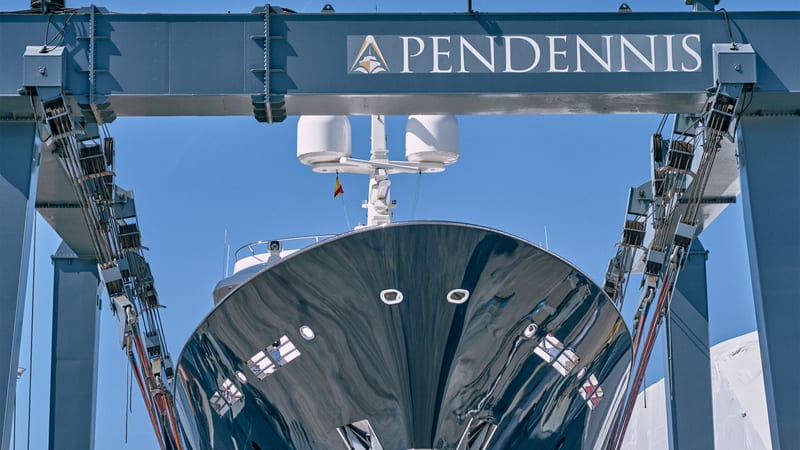 Superyacht bow suspended in Pendennis Vilanova's lifting dock beneath large blue gantry crane bearing the Pendennis logo.