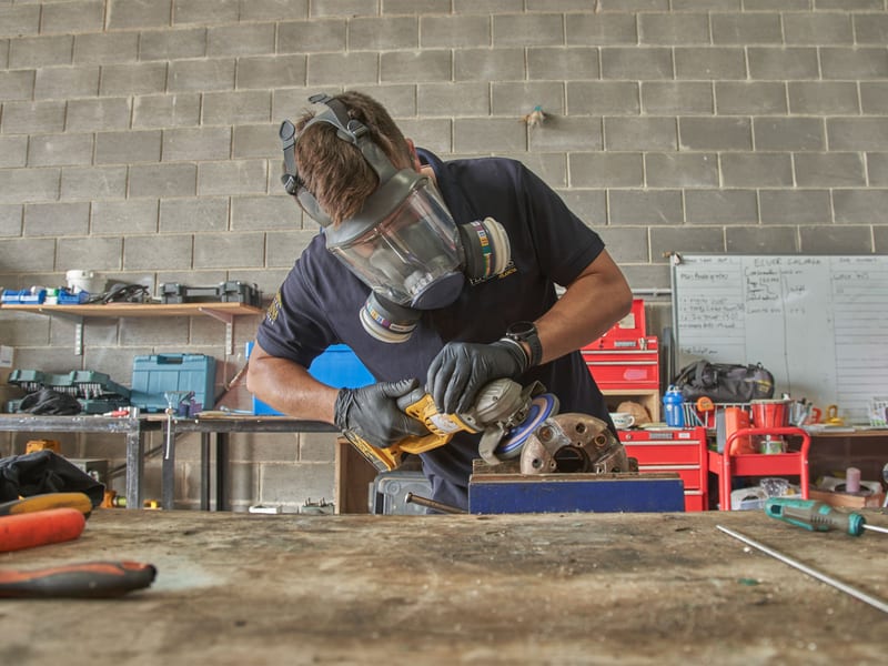 Pendennis Vilanova technician wearing a respirator and gloves while grinding a metal part in the shipyard workshop.