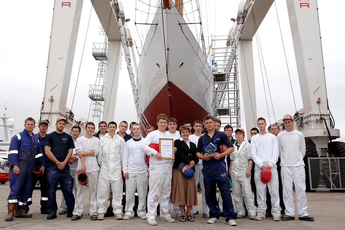 Pendennis’s first group of Surface Finishing apprentices pose in front of a yacht in the travel hoist, holding certificates and awards.