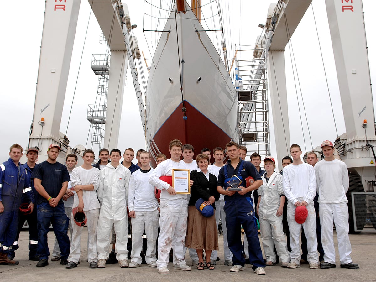 Pendennis’s first group of Surface Finishing apprentices pose in front of a yacht in the travel hoist, holding certificates and awards.